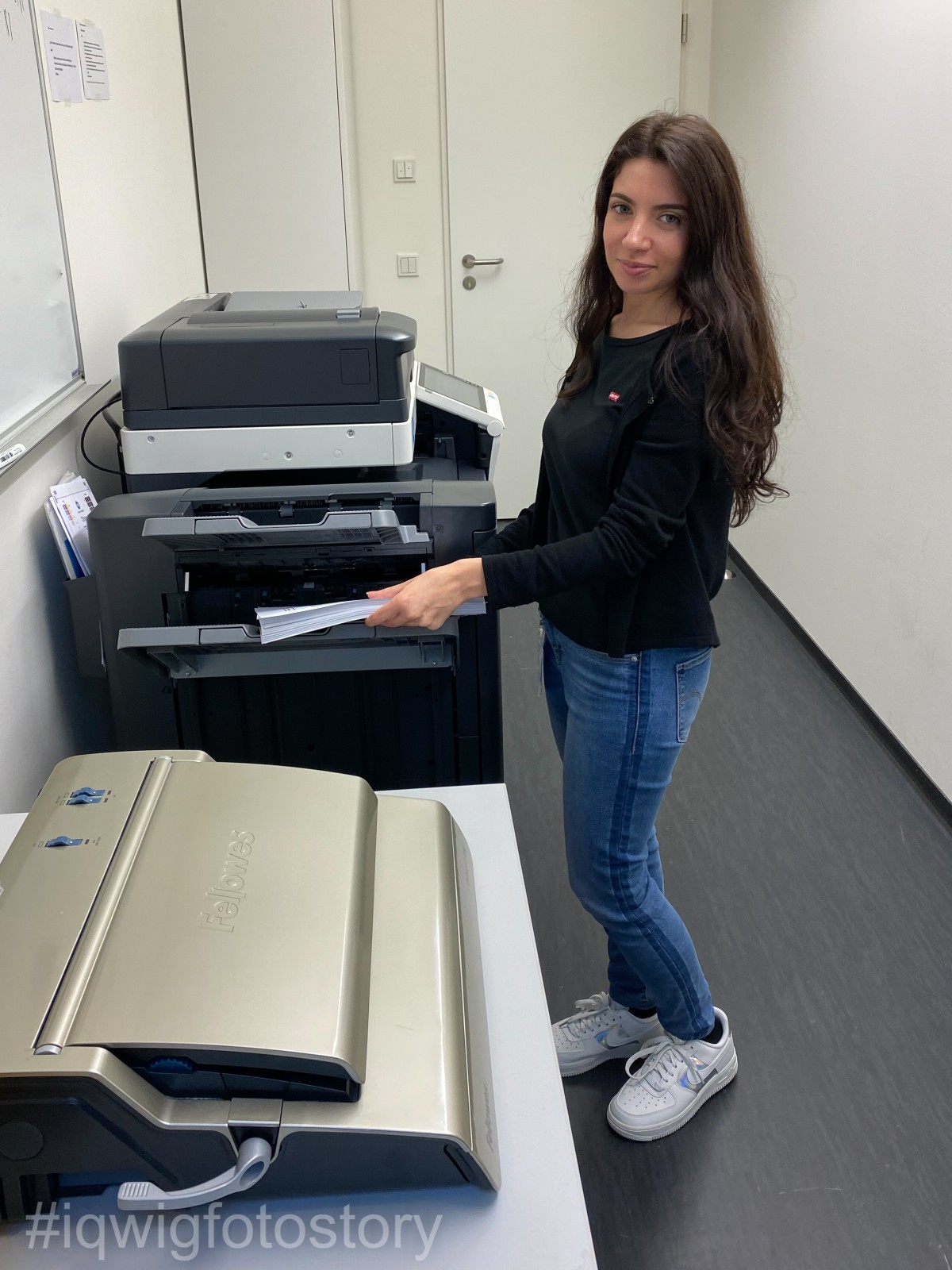 A woman is taking a stack of documents out of a printer and is smiling at the camera. She has long hair and is wearing a black top, blue jeans and white trainers.