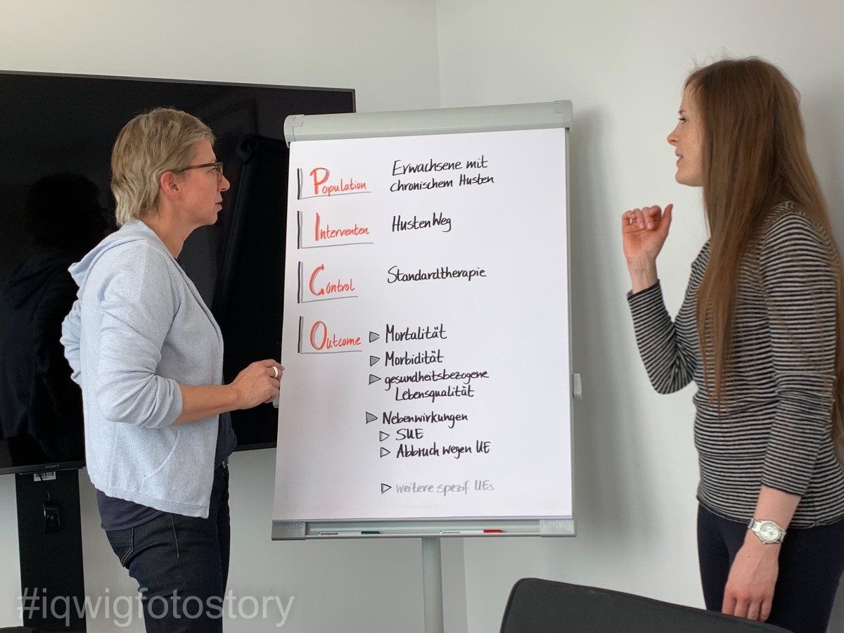 Two women stand facing each other. They look intently at a flipchart standing between them. On it is the PICO scheme: Population, Intervention, Control and Outcome for the studies on the cough medicine "HustenWeg". The woman on the right has long hair and is wearing a striped top. The woman on the left has short hair, and is wearing glasses and a white top.