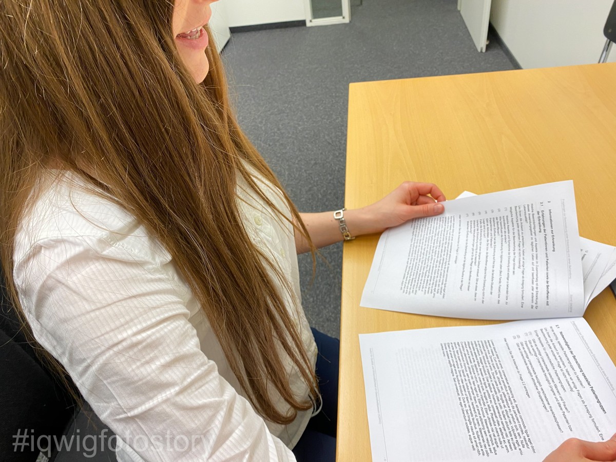 A woman is sitting at a table, reading a document that she is holding in her hands. She has long hair and is wearing a white shirt.