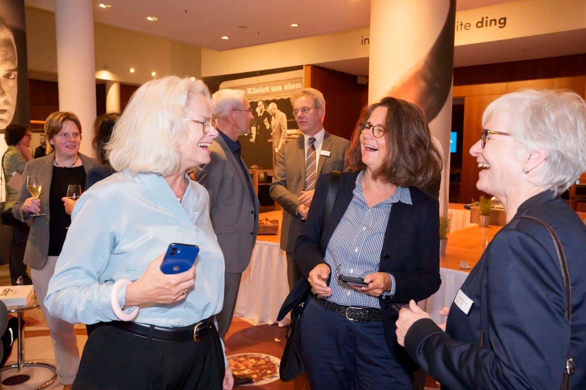 Kirsten Kappert-Gonther (MdB), Mirjam Mann (ACHSE e. V.) und Andrea Benecke (BPtK) im Gespräch. Im Hintergrund Stefan Sauerland aus dem IQWiG mit Stefan Gronemeyer (Medizinischer Dienst Bund) und Teile der Kunstausstellung in der Landesvertretung.