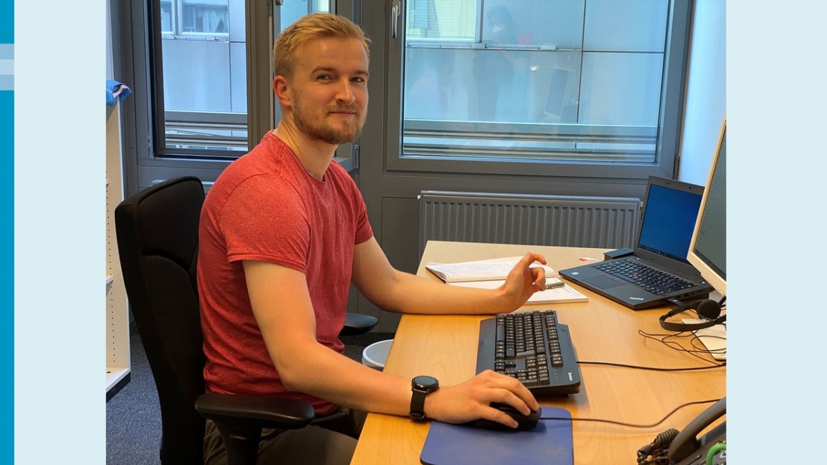 Ein junger Mann mit blondem Haar und Bart sitzt an einem Schreibtisch. Er trägt ein rotes T-Shirt und eine Armbanduhr und blickt freundlich in die Kamera. Vor ihm eine Tastatur, ein blaues Mauspad, ein Flachbildschirm, ein Headset und ein Laptop. Im Hintergrund eine graue Heizung und eine graue Wand mit Fenstern.