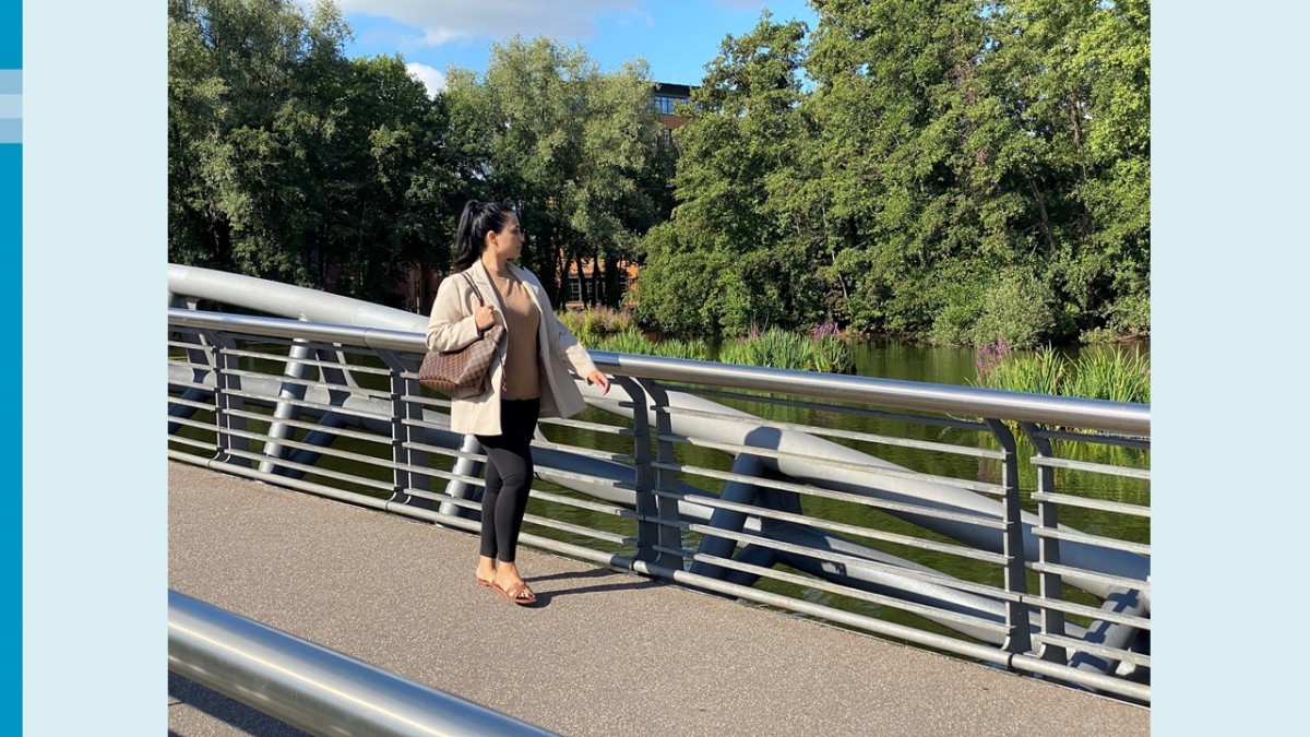 Eine Frau mit dunklem Haar und Pferdeschwanz, beigem Oberteil, hellem Sakko, schwarzer Hose und Sandalen läuft von links nach rechts über eine Fußgängerbrücke. Sie hat eine Umhängetasche über der rechten Schulter. Hinter ihr ein See, Bäume und etwas blauer Himmel.