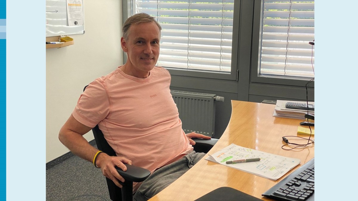 Ein Mann in einem rosa T-Shirt sitzt entspannt auf einem Bürostuhl und lächelt in die Kamera. Vor ihm ein Schreibtisch mit einer Ausbuchtung, hinter ihm ein Stück von einem Whiteboard und zwei Fenster mit heruntergefahrenen Jalousien.