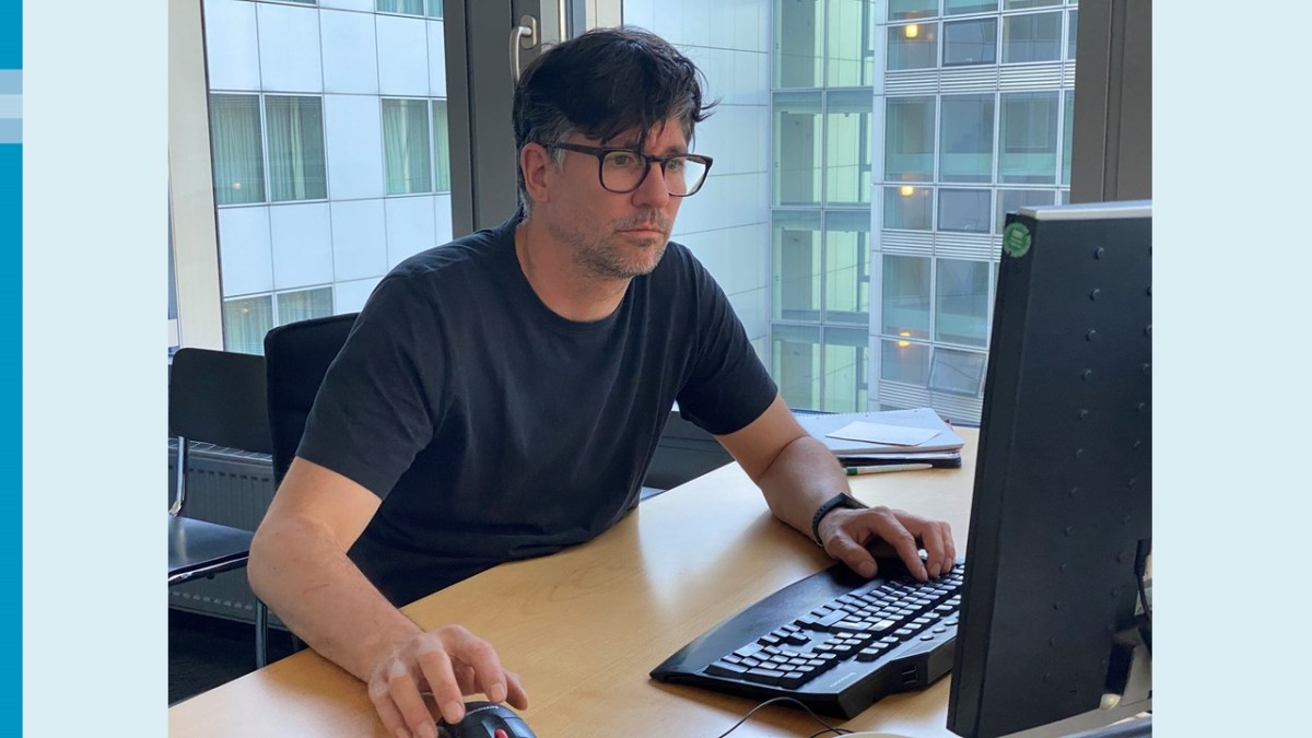 Ein Mann mit dunklem Haar und dunkel gerahmter Brille sitzt in einem Büro und arbeitet konzentriert am Computer. Im Hintergrund zwei Fenster, durch die man eine graue Hotelfassade sieht.