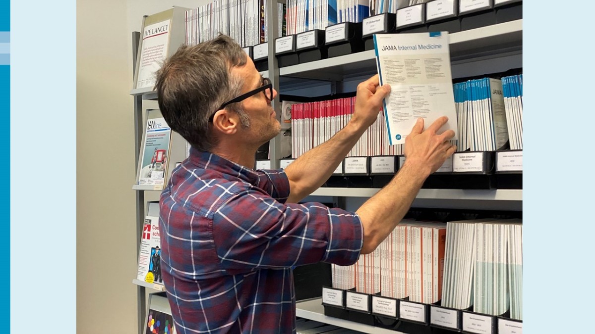 Ein Mann mit dunklem Haar, grauen Schläfen und einer Brille mit dunklem Gestell steht vor einem Zeitschriftenregal in der Bibliothek. Er hat ein großkariertes Hemd mit hochgekrempelten Ärmeln an. Er zieht gerade eine Ausgabe der Zeitschrift "JAMA Internal Medicine" aus einem Schuber.