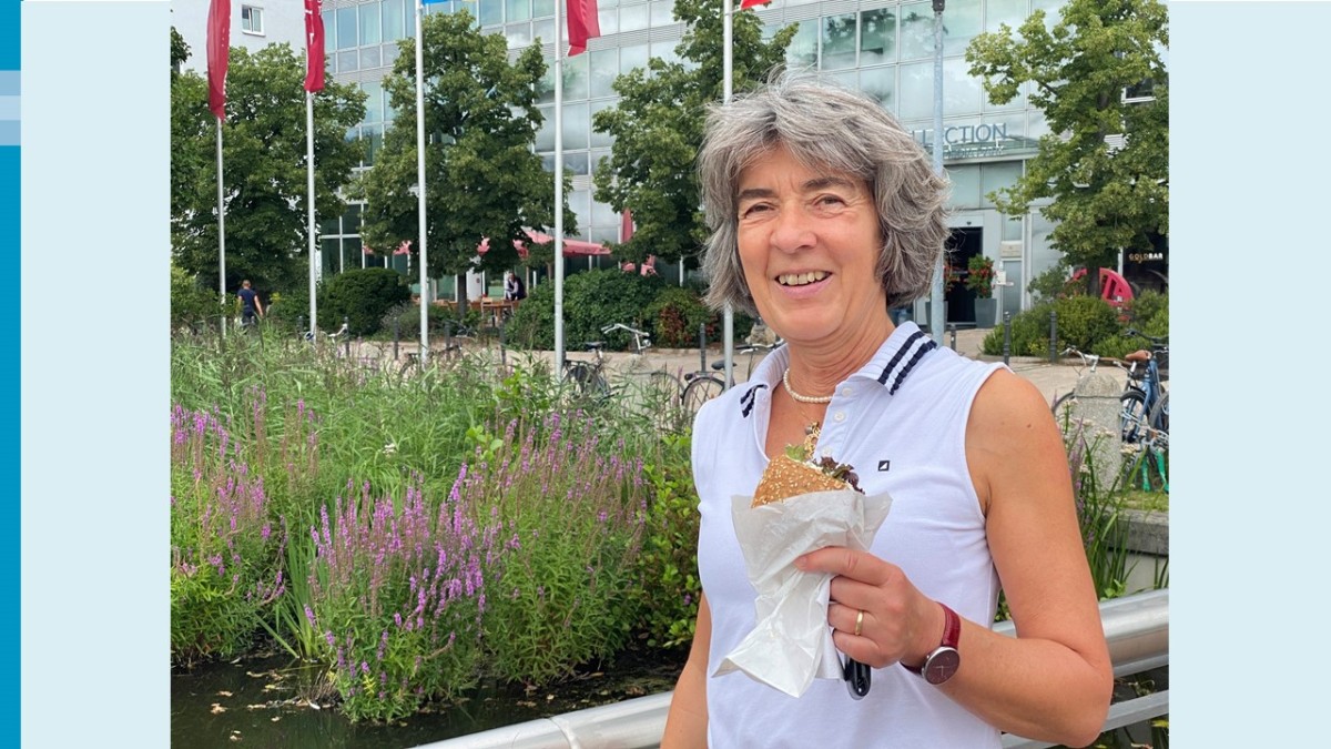 Eine gut gelaunte Frau mit weißem Oberteil hält ein belegtes Brötchen in der Hand. Im Hintergrund befinden sich neben einem Gebüsch und Bäumen, ein großes Gebäude mit Glasfassade.