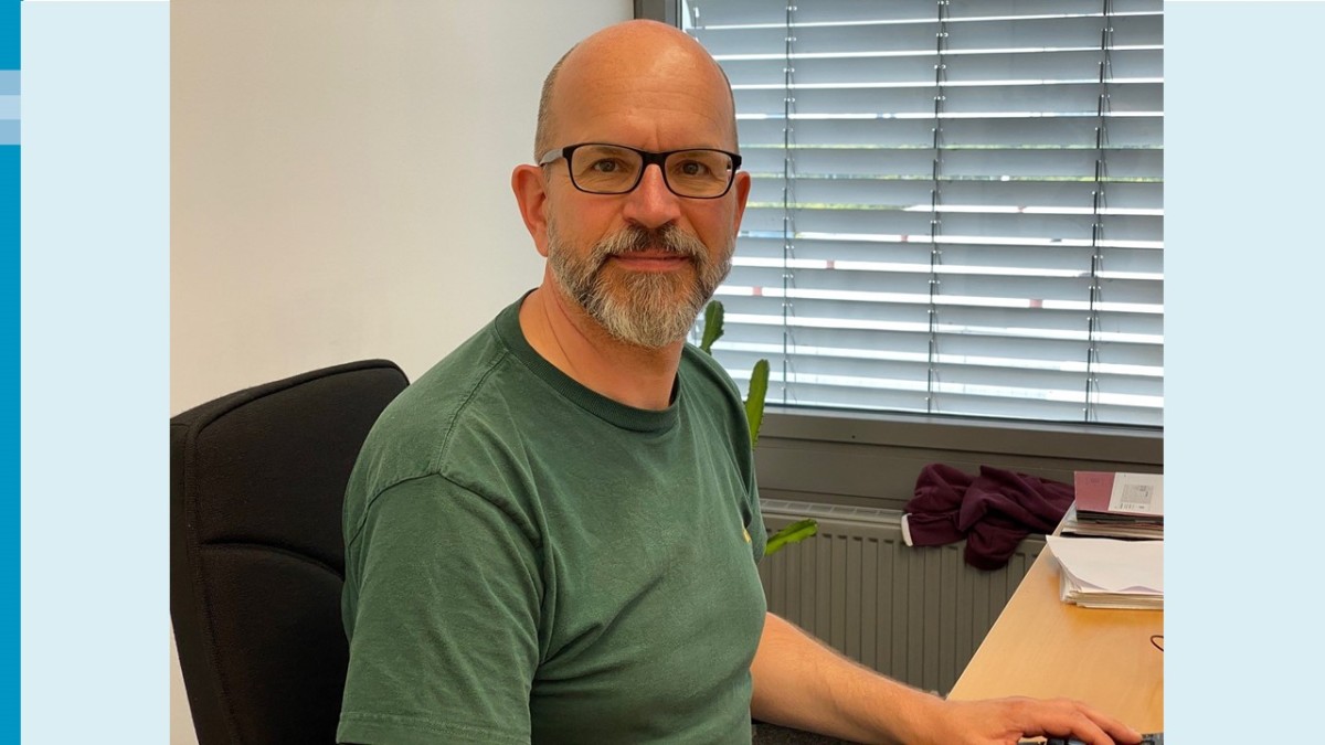 Ein Mann mit Bart und dunkler Brille schaut freundlich in die Kamera. Er sitzt auf einem schwarzen Bürostuhl mit halblanger Stuhllehne. Die linke Hand bedient eine Computer-Tastatur. Im Hintergrund befindet sich ein Fenster mit geschlossenen Jalousien. Unterhalb des Fensters ist ein Heizkörper zu sehen; auf diesem liegt ein roter Pullover. Auf der linken Seite des Schreibtisches liegen Dokumente. Links neben dem Mann ist außerdem ein Zimmerkaktus zu sehen.