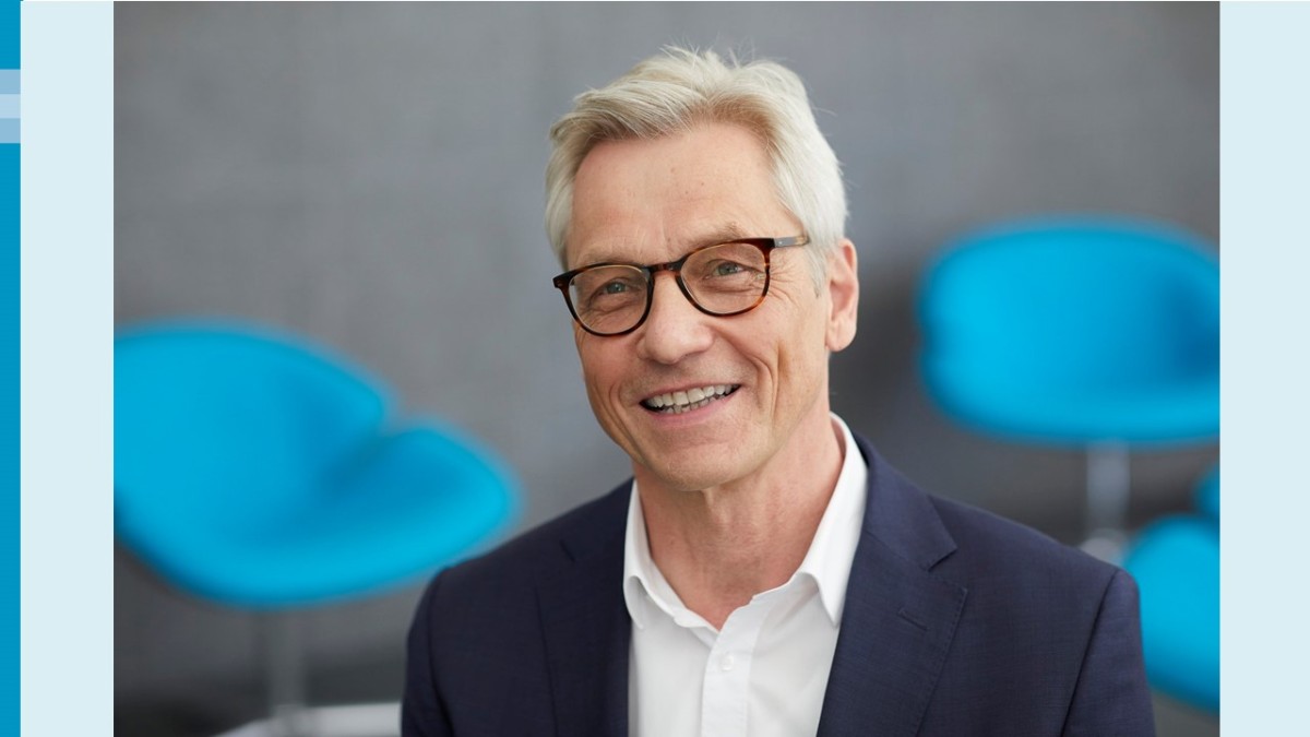 Portraitfoto eines Mannes mit hellgrauem Haar und dunkler Brille, der freundlich in die Kamera schaut. Er trägt ein weißes Hemd mit einem dunkelbauen Jackett darüber. Im Hintergrund sind unscharf zwei blaue Stühle und eine graue Wand zu sehen.