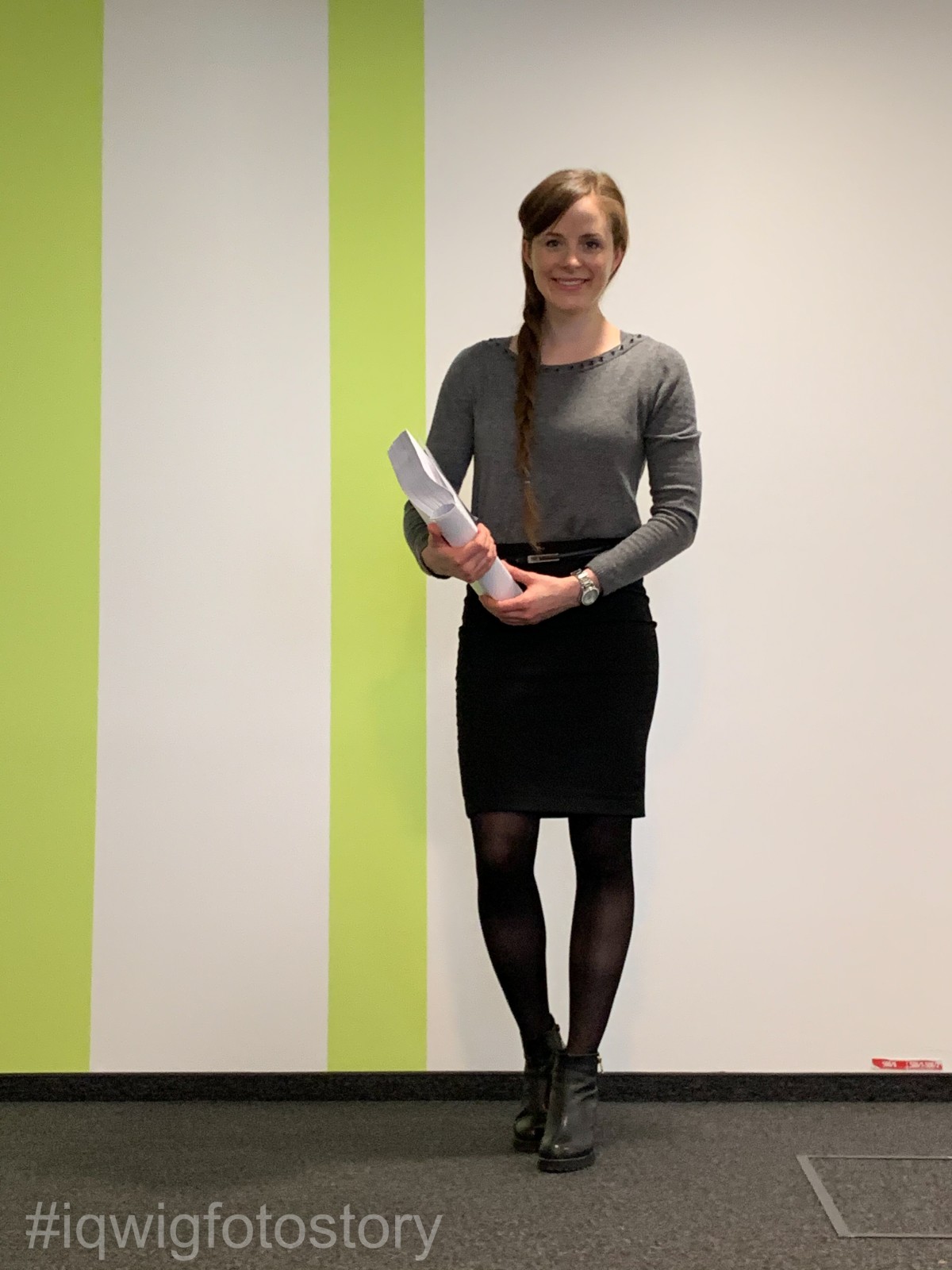 A woman in a grey pullover, black skirt and black ankle boots looks happily into the camera. Her long brown hair is braided. She holds a thick document in her arms. Behind her is a white corridor wall with two vertical light green stripes.