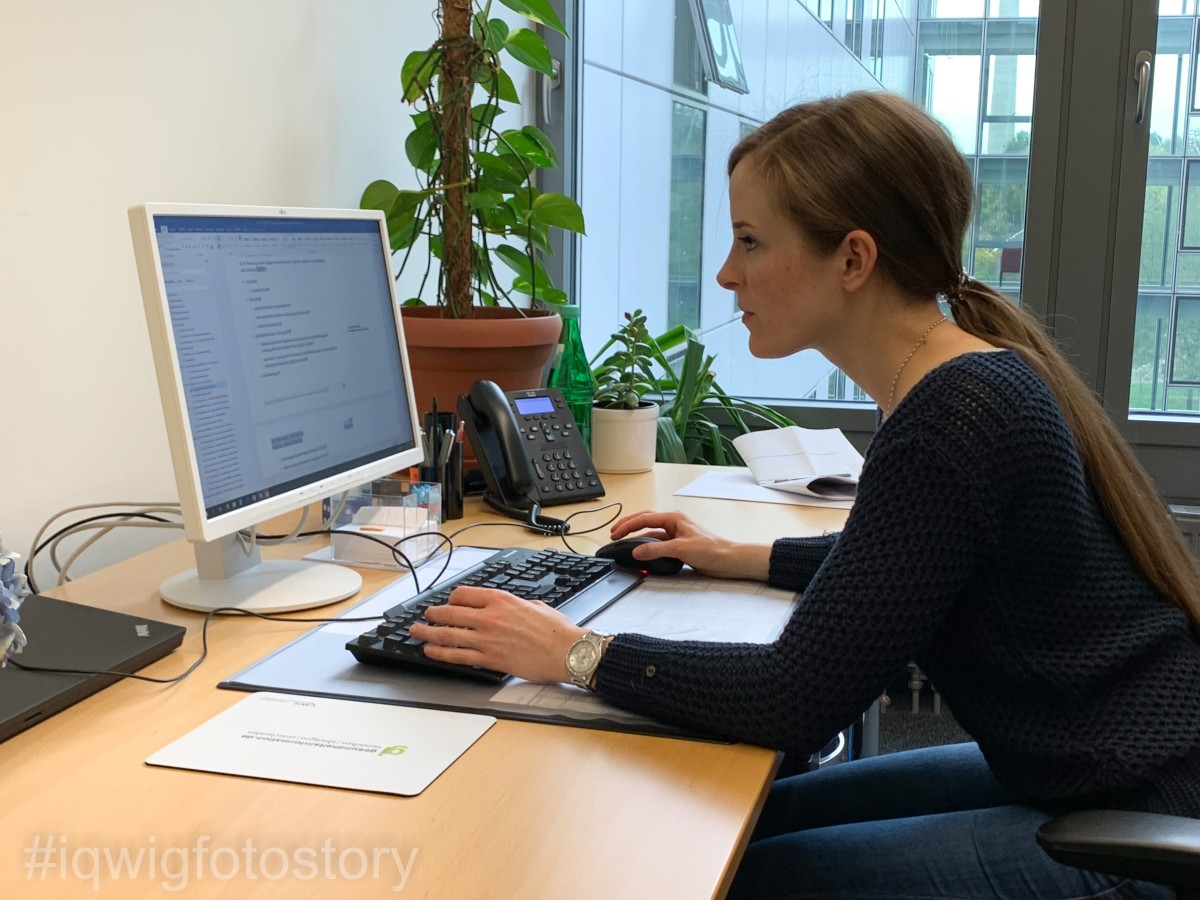 A woman is sitting at her desk, looking intently at the computer screen in front of her. She has her hair in a braid and is wearing a black top and blue jeans. Her right hand is controlling the computer mouse. Her left hand is typing on the computer keyboard. There are writing utensils and a telephone on the desk. In the background, there are plants and a window.