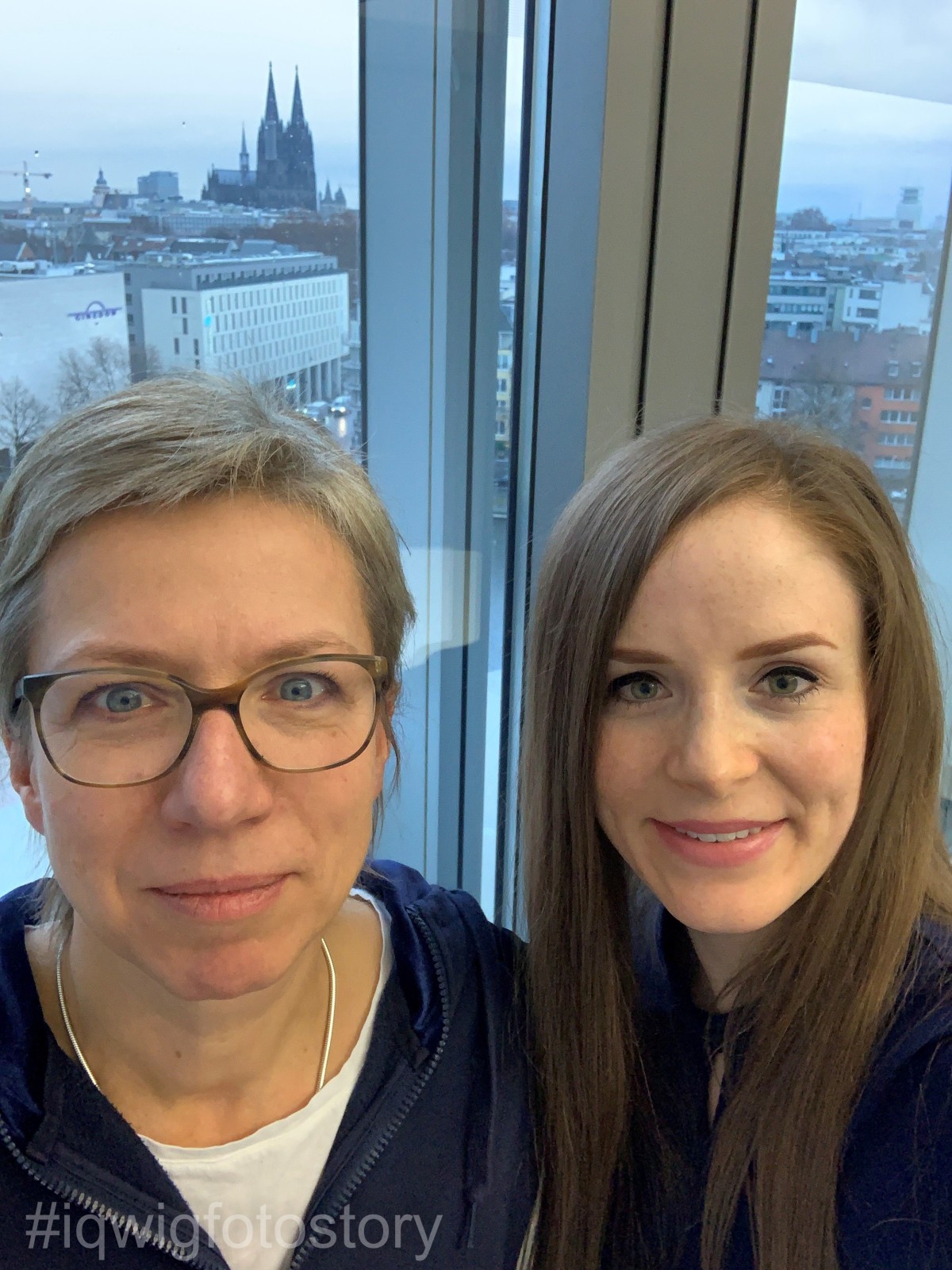 Two women are smiling at the camera. The woman on the left has short hair and is wearing glasses. The woman on the right has long hair. Cologne Cathedral can be seen through the window behind them.