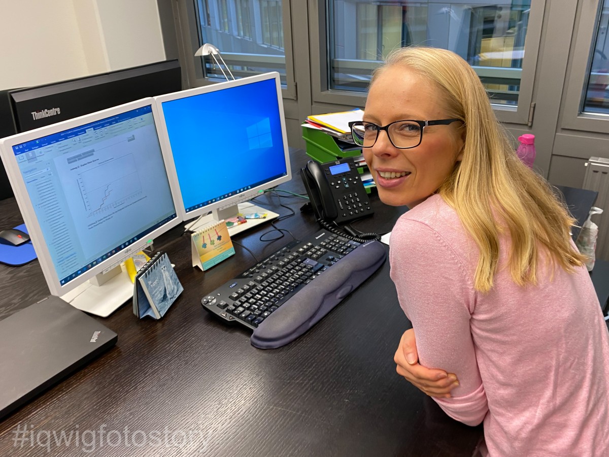 A woman is sitting comfortably at her desk, smiling into the camera. She has long hair, and is wearing glasses and a pink top. In front of her on the desk are two screens, a computer keyboard, a laptop, a telephone and documents. In the background to the right are windows.