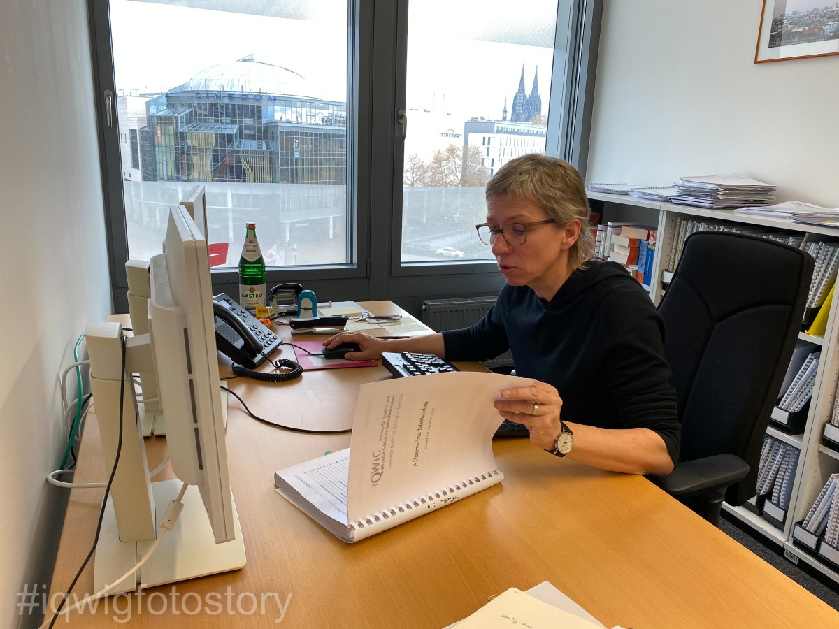 A woman with short hair, who is wearing glasses and a black top, is sitting at her desk, looking at a report. She is wearing a watch on her left wrist. She is leafing through the report with her left hand. Her right hand is controlling the computer mouse. In front of her on the desk are a computer keyboard, two screens and a telephone. To the right of the telephone there are more writing utensils and a water bottle. Behind the woman is a shelf with several bound reports and books. In the background, the cathedral can be seen through a window.