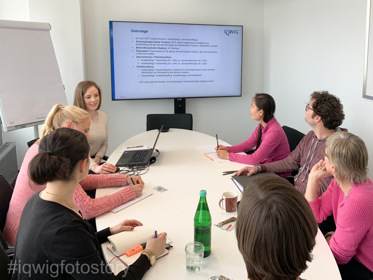 Eine Gruppe Menschen sitzt um einen ovalen Tisch und schaut interessiert auf einen großen Bildschirm mit Informationen zur Datenlage. Auf der linken Seite befindet sich ein Flipchart.