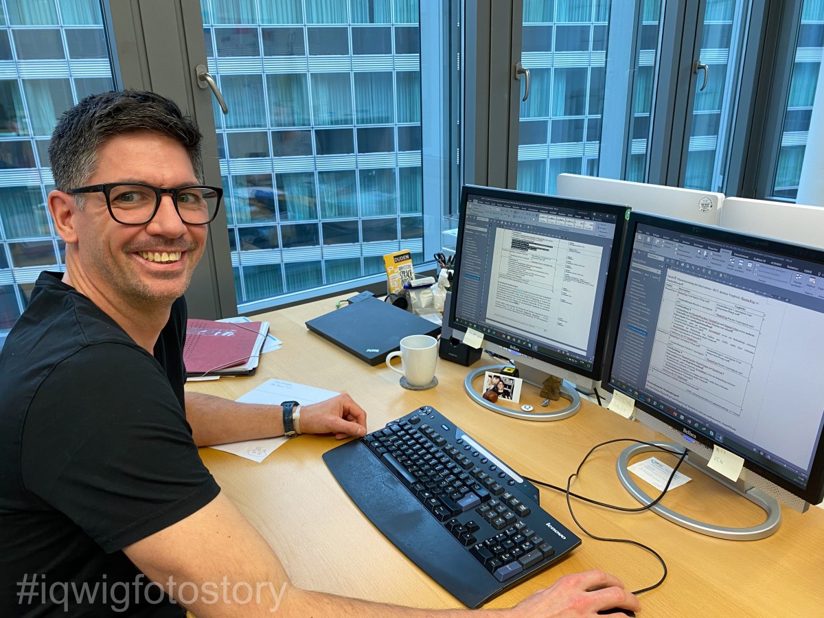A man with short dark hair, who is wearing glasses and a black T-shirt, is sitting at a desk and smiling at the camera. MS Word files can be seen on the two screens in front of him. In the background is a window, through which the building opposite can be seen.