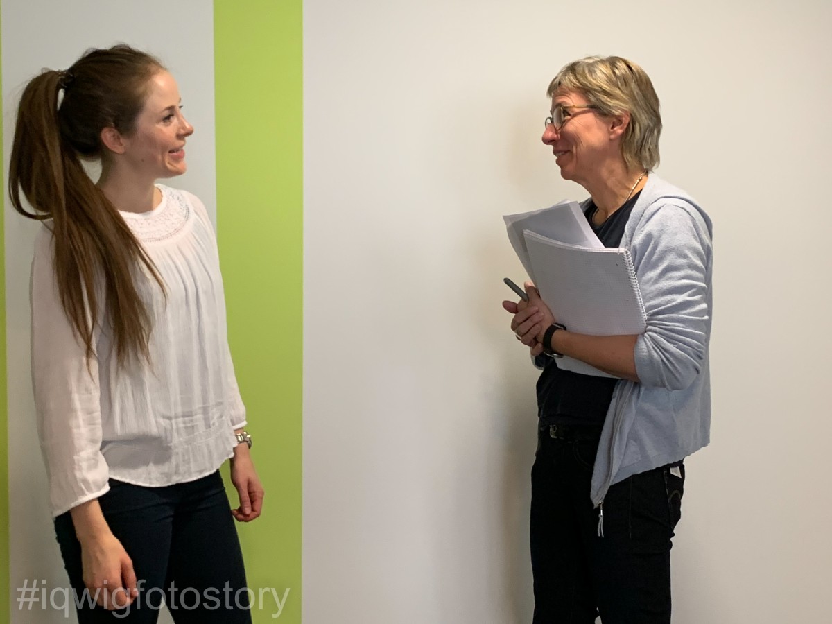 Two women are standing in front of a white wall with a light green vertical stripe. The woman on the right is holding two large documents and a pen. They are discussing a problem and seem relieved to have solved it.