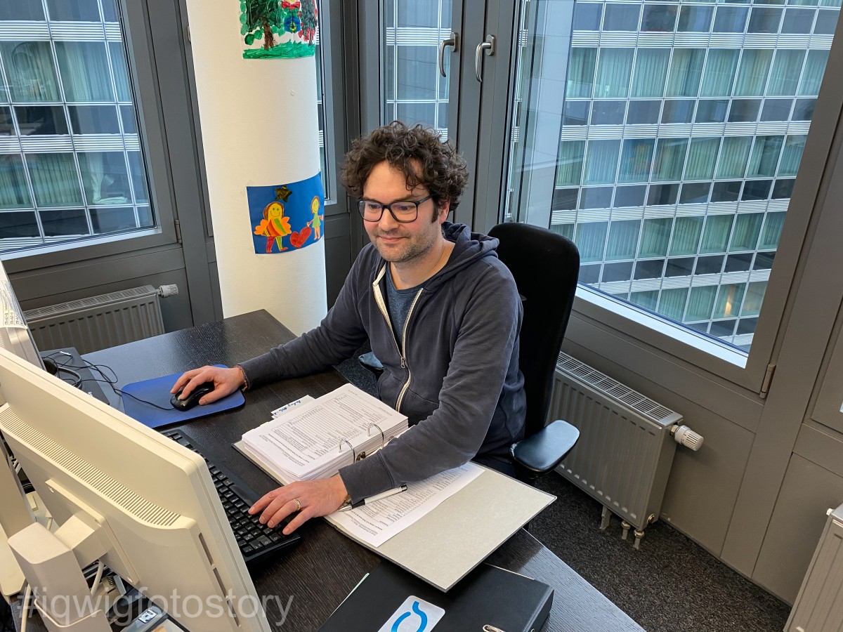 A man is sitting at a desk, working on a computer. He is smiling and looking intently at the screen. He has brown curly hair, and is wearing glasses and a grey sweatshirt. There are two folders of documents on the table. In the background are windows. Through them, you can see the facade of the building opposite.