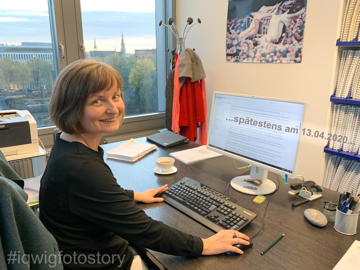 A woman is sitting at a desk, and turns towards the camera and smiles. She has medium-length hair and is wearing a black top. There is a computer keyboard and screen on the desk. Also on the desk are a cup of tea, a writing pad, a jar of pens, a stapler and a puncher. To the left of the woman is a window. 