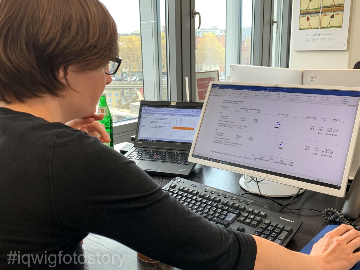 A woman is sitting at her desk, absorbed in her work on the computer. She has medium-length brown hair, and is wearing glasses and a black top. On the desk in front of her is a computer keyboard and screen. To the left, there is an open laptop and a water bottle. There is a window in the background on the left.