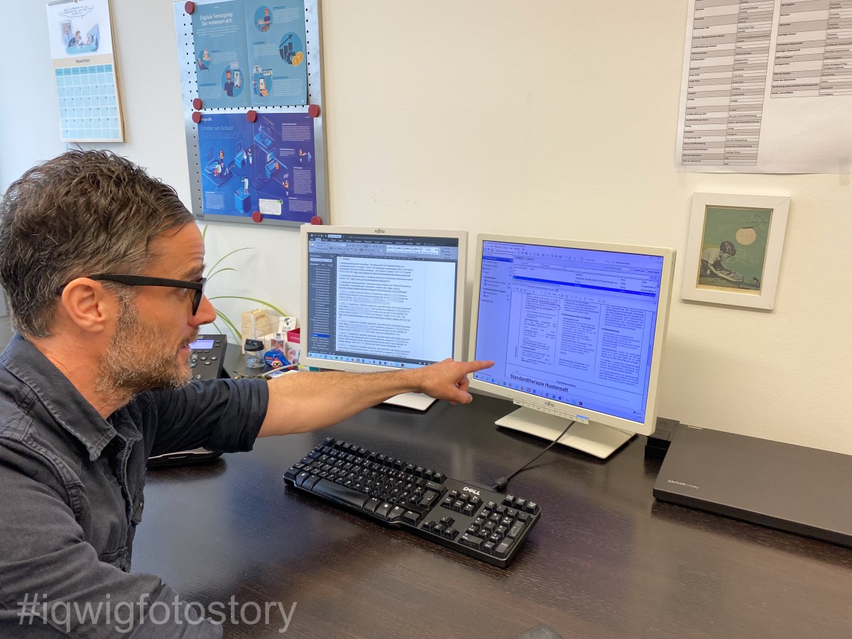 A man is sitting at his desk, pointing with his left index finger at the screen in front of him with a look of surprise. On the desk in front of him are a computer keyboard, two screens and a laptop. On the wall in front of him are two posters, a table, a picture and a calendar.