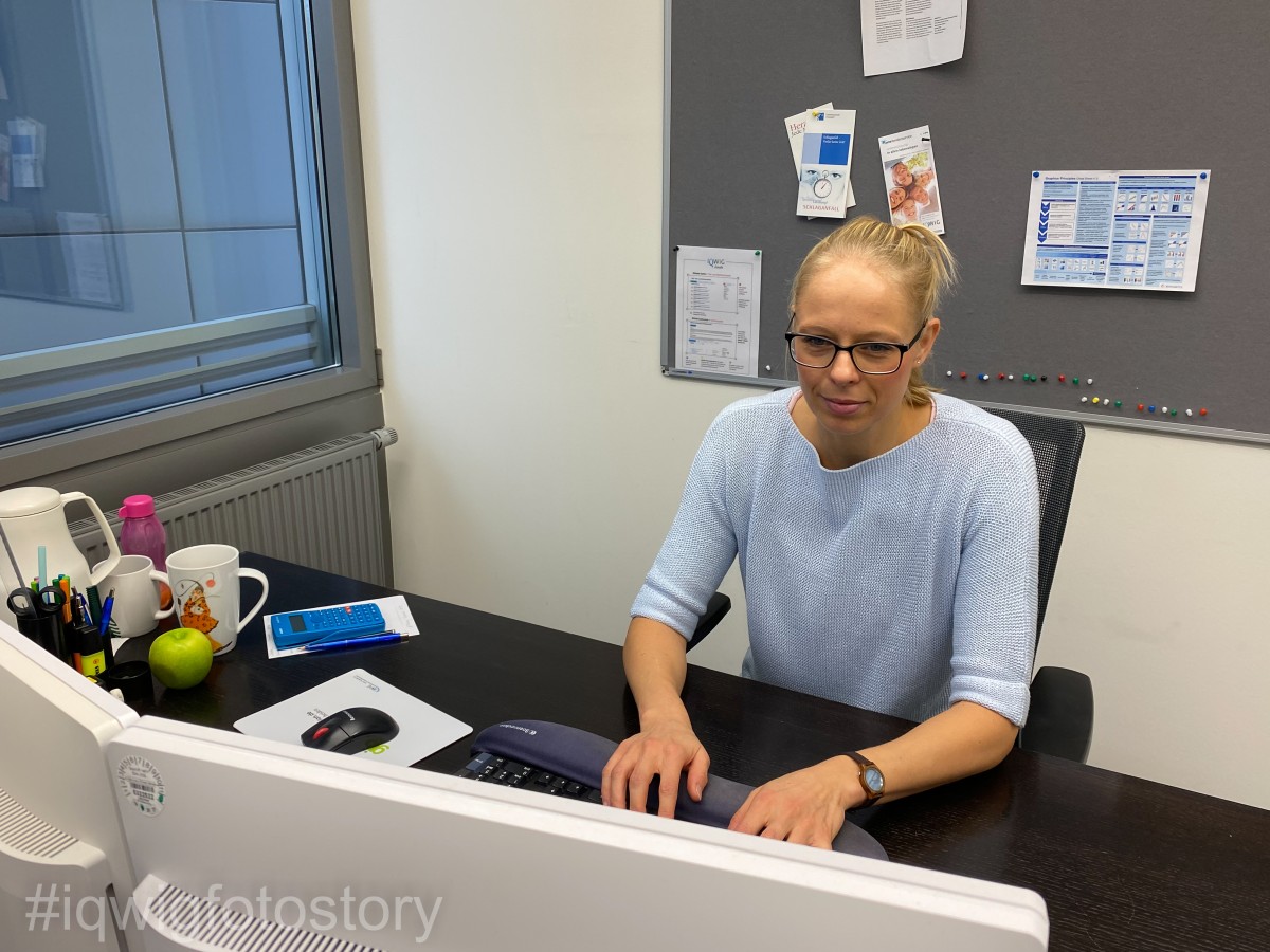 Eine Frau sitzt an ihrem Schreibtisch und arbeitet am Computer. Sie schaut konzentriert auf den Monitor und trägt ihre Haare zum Zopf, eine Brille und ein hellblaues Oberteil. Vor ihr auf dem Schreibtisch stehen zwei Monitore, eine Computer-Tastatur und eine Computer-Maus. Auf der linken Seite des Schreibtisches liegen Schreibutensilien. Außerdem stehen dort zwei Tassen eine Thermoskanne und ein Apfel.