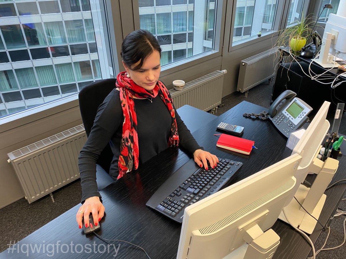 Eine Frau sitzt am Schreibtisch und arbeitet am Computer. Sie schaut konzentriert auf den Monitor. Sie trägt die Haare zum Zopf, ein schwarzes Oberteil und einen rot-schwarzen Schal. Auf dem Schreibtisch befinden sich auf der rechten Seite ein Notizbuch, Schreibutensilien und ein Telefon. Hinter der Frau ist ein Fenster. Durch dieses ist die Fassade des Gebäudes gegenüber zu sehen.