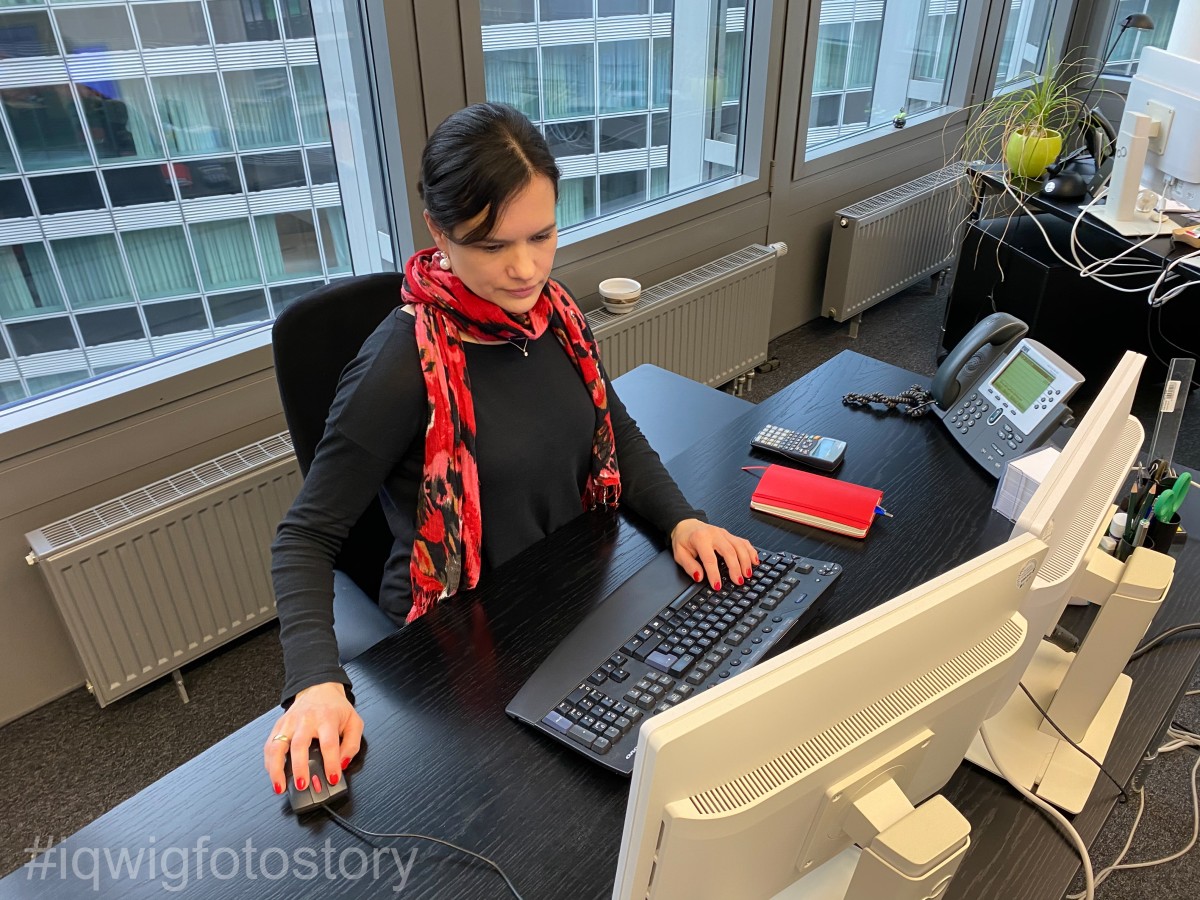 A woman is sitting at a desk, working on a computer. She is looking intently at the screen. She has braided hair and is wearing a black top and a red and black scarf. On the desk, to the right, there is a notepad, writing utensils and a telephone. Behind the woman is a window. The facade of the building opposite can be seen through the window.