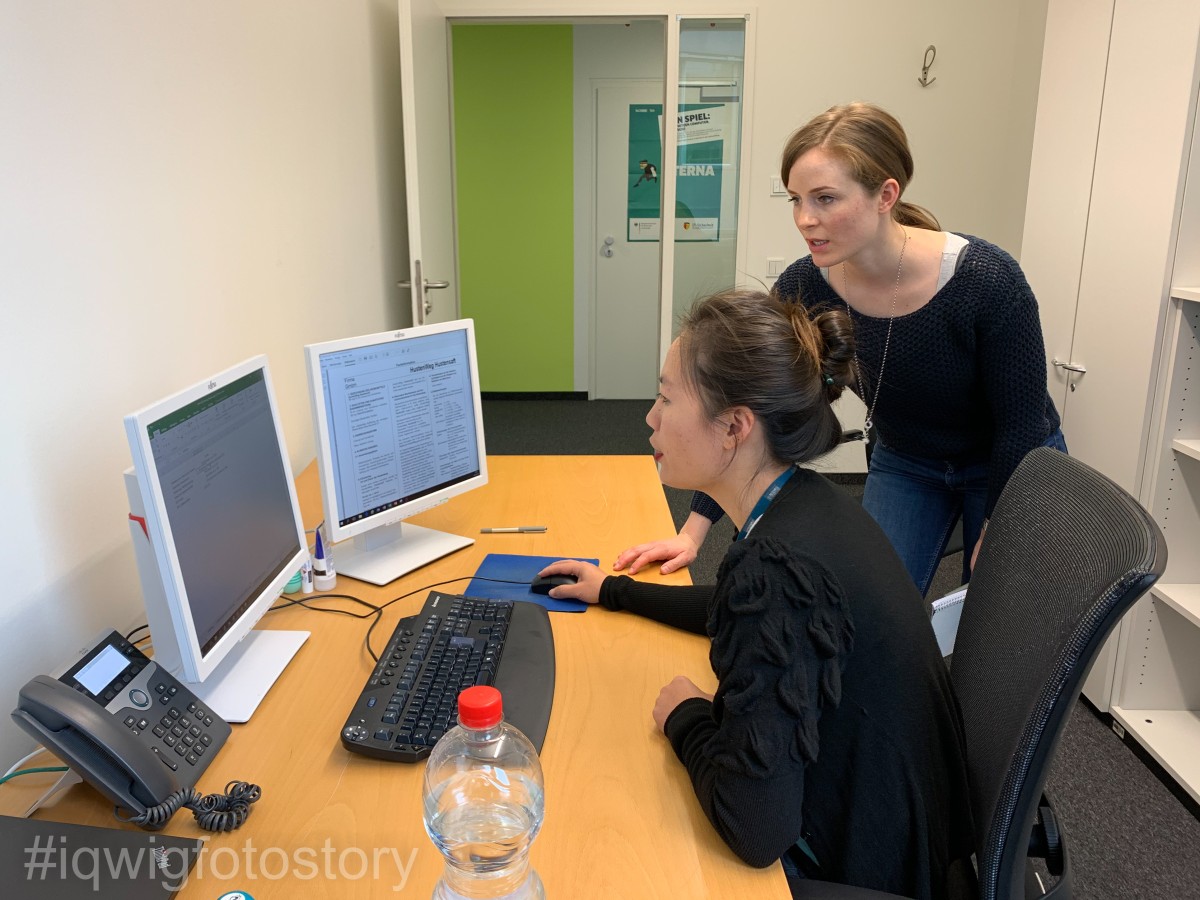 Two women are looking at a computer screen on a desk, with a look of surprise. The woman in the foreground is sitting in a chair, using a computer mouse with her right hand. She has her hair in a braid and is wearing a black top. To her right is the other woman. She leans forward with her upper body towards the computer screen and leans on the table with her right hand. She has her hair in a braid and is wearing a black top and blue jeans. There is a water bottle, a second screen and a telephone on the desk. The computer keyboard is in front of the seated woman.