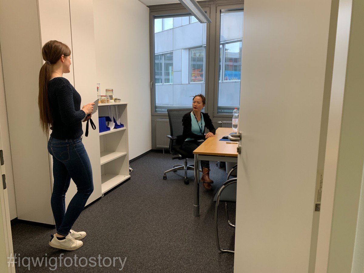 Eine Frau steht in einem Büro. Sie trägt lange Haare zum Zopf, ein dunkles Oberteil, eine blaue Jeans und weiße Schuhe. Sie unterhält sich mit einer anderen Frau, die in der Mitte des Büros an einem Schreibtisch sitzt. Sie trägt die Haare zum Zopf und eine schwarze Sweatshirtjacke über ein türkisfarbenes Oberteil. Um den Hals trägt sie ein Schlüsselband. Im Büro befinden sich auf der linken Seite ein Aktenschrank und ein Regalelement. Im Hintergrund ist ein Fenster. Durch dieses ist die Fassade des Gebäudes gegenüber zu sehen.