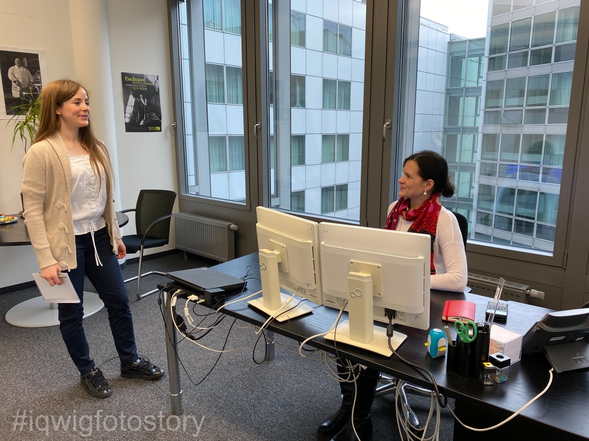 Two women are talking in an office with large windows in the background. The woman on the right is sitting at a desk. There are two screens, a laptop, writing utensils and a telephone. She has her hair in a braid and is wearing a white top and a red and black scarf. She is looking at the other woman in an attentive and friendly way. The woman on the left is standing to the right of the desk and looking at her colleague. She has long hair and is wearing a beige cardigan over a white shirt, blue jeans and black shoes. She is holding a notepad and pen in her right hand.