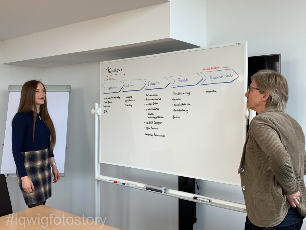 Two women are facing each other. There is a magnetic board between them. A project protocol is drawn on it. The woman on the right has short hair, and is wearing glasses and a light brown corduroy jacket. The woman on the left has long hair, and is wearing a dark blue top and a checked skirt.