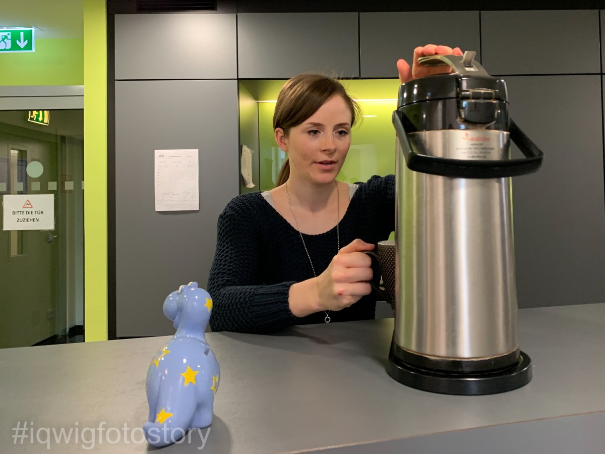 A woman is standing at a counter next to a coffee pot, filling her cup with coffee. The woman has her hair in a braid, and is wearing a black top and necklace. The pot is on a counter. On the left side of the counter is a small porcelain dinosaur with a coin slot. There is a grey cupboard in the background and a glass door on the left.