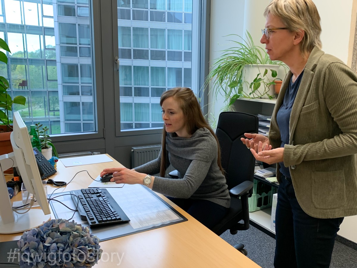 Eine langhaarige Frau in einem grauen Pullover sitzt an einem Schreibtisch. Neben ihr steht eine kurzhaarige Frau in einer Cordjacke. Sie hat beide Handflächen nach oben gedreht, eine Geste, die Ratlosigkeit ausdrückt. Beide schauen auf einen Flachbildschirm. Im Hintergrund Fenster und Grünpflanzen.