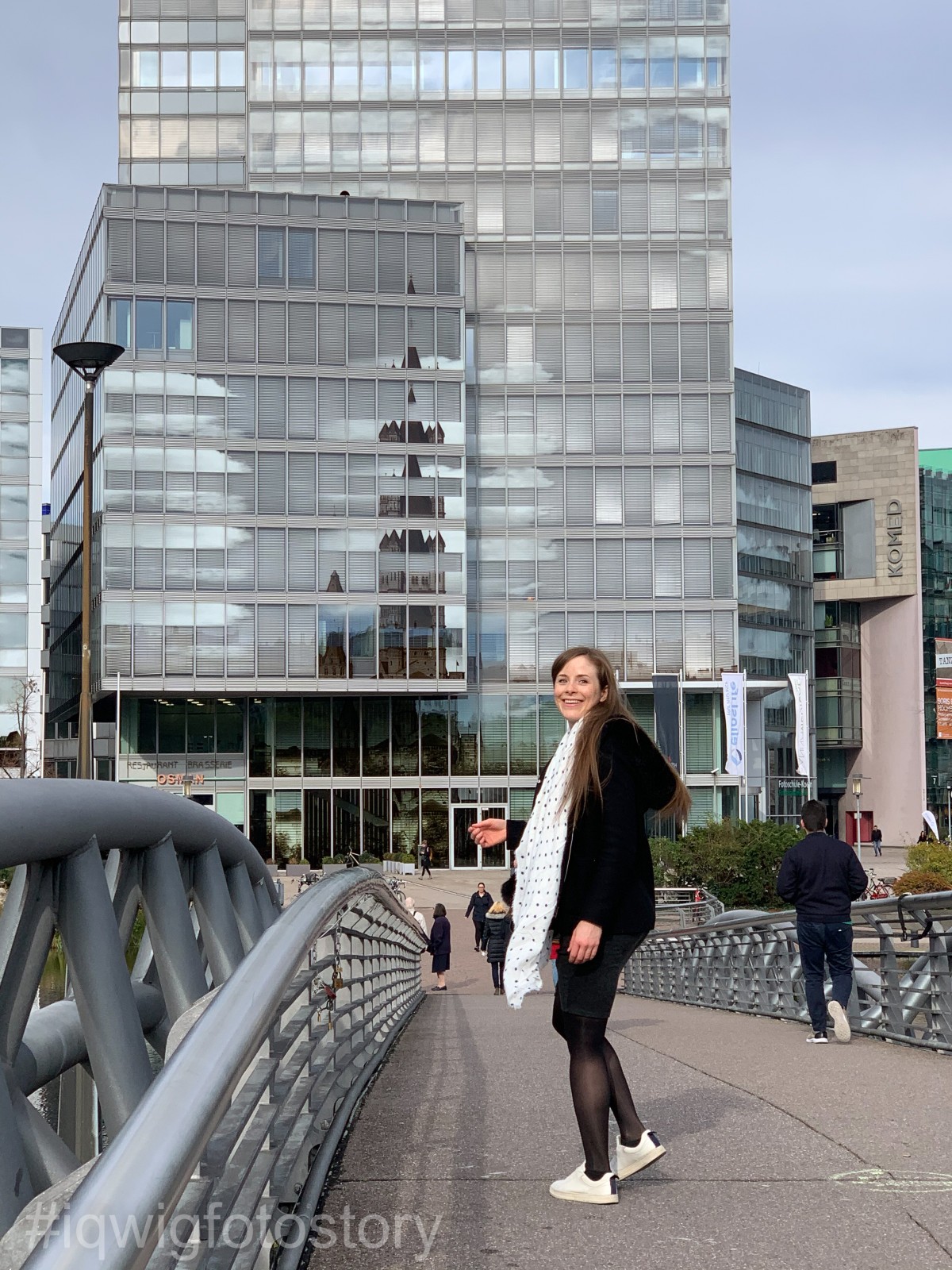 IQWiG staff member Vanessa is standing on a bridge, smiling into the camera. In the background is a high-rise building with a glass facade.