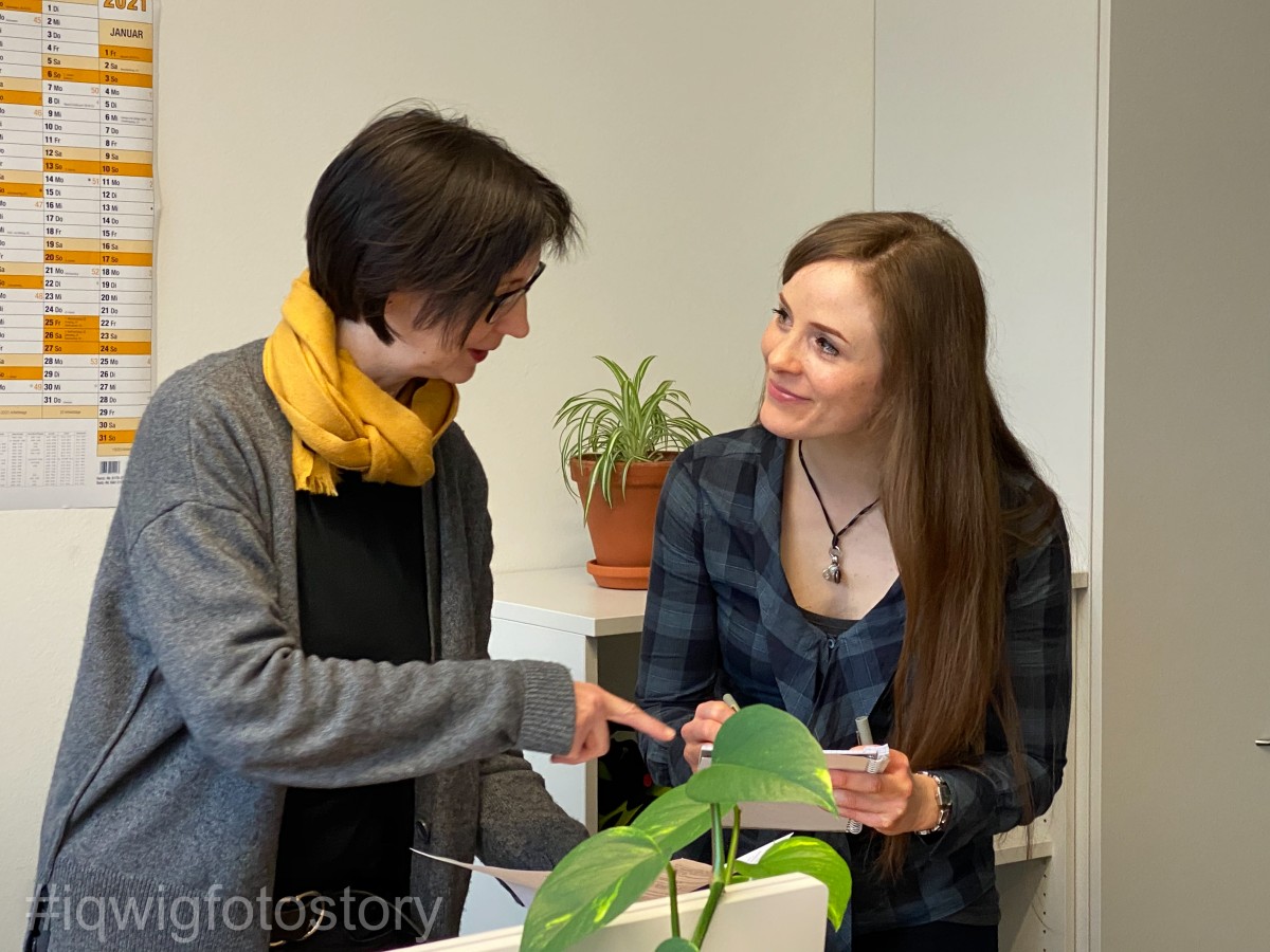 Two women are standing next to each other, discussing a document. The woman on the right has long hair and is wearing a checked shirt. She is holding the document. The woman on the left has chin-length hair, and is wearing glasses, a grey cardigan with a black sweater underneath, and a yellow scarf. There is a plant in front of each of the women and in the background.