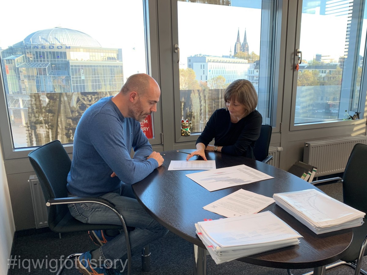 A man and a woman are sitting opposite each other at a table. They are studying a document lying between them. The man has short hair, a beard and a blue shirt. The woman has medium-length hair and is wearing a black top. There are two other single documents and two stacks of documents on the table. Behind them, on the left, is a cinema with a dome and, on the right, Cologne Cathedral.