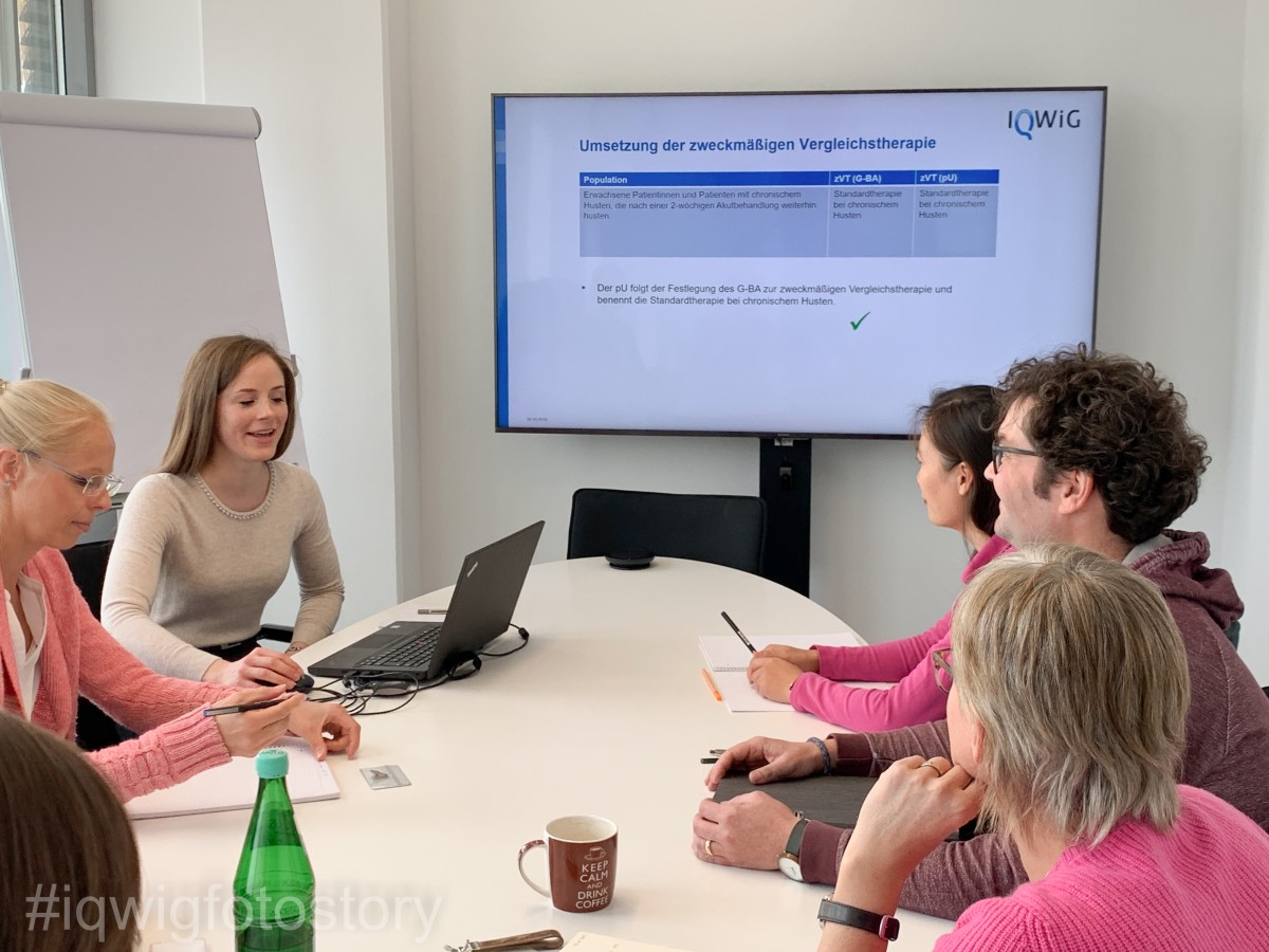 A group of people are sitting around an oval table, intently following a presentation. A woman in the group has a laptop in front of her and is giving the presentation. Behind her is a flipchart. There is a large screen in the middle and further back.