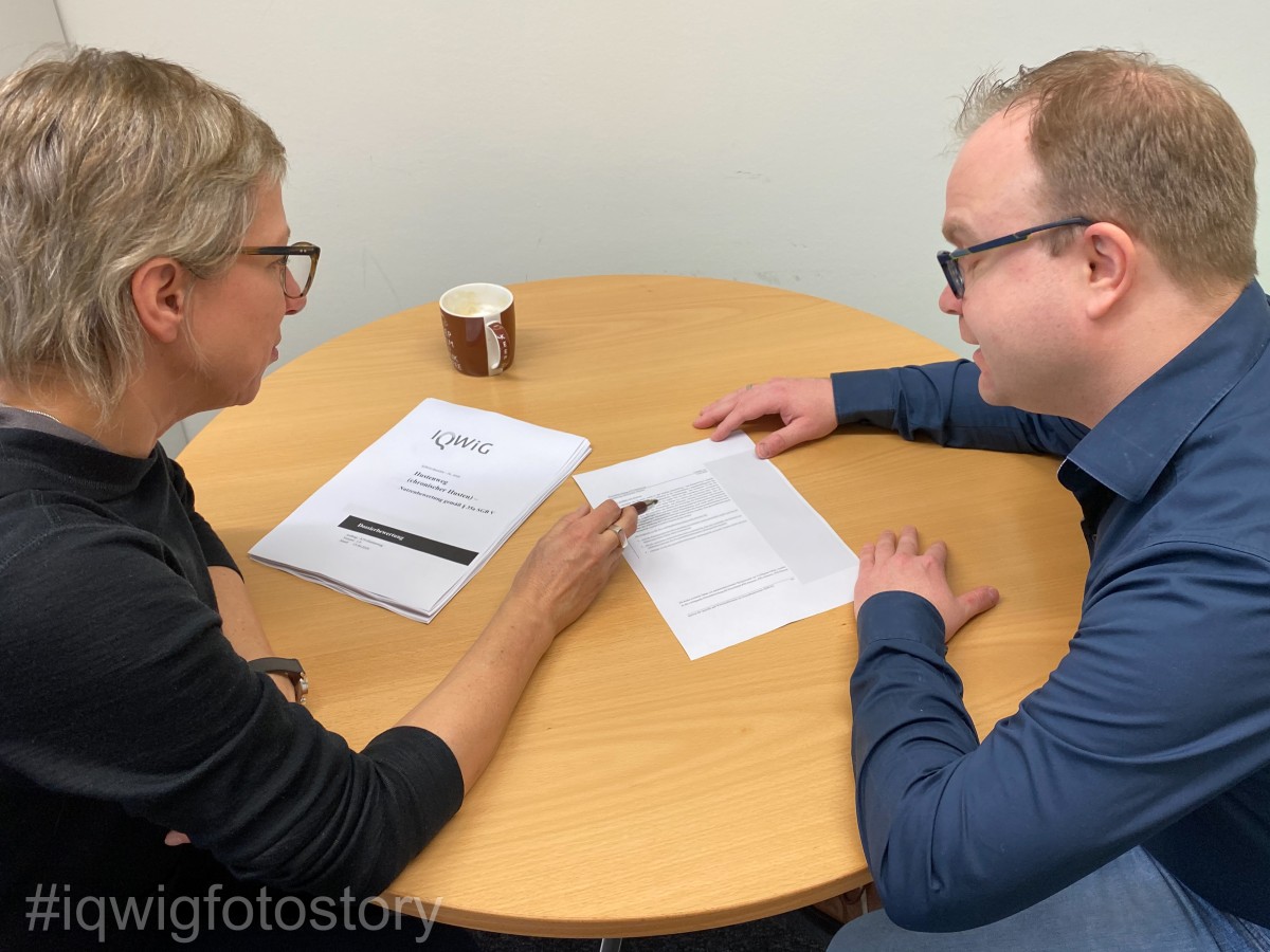 A man and a woman are sitting at a round table looking at a document. The woman has short hair, and is wearing glasses and a black sweatshirt. The man has short hair, and is wearing glasses and a blue shirt. There is another pile of documents on the table and a coffee cup.