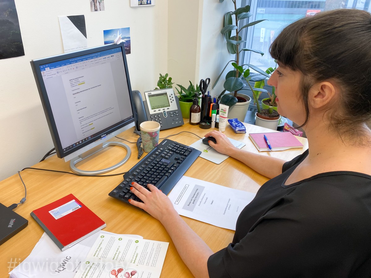 A woman with dark hair in a bun and wearing a black top is sitting at a desk, working on a text. In front of her is a leaflet with a medical illustration and a red notepad. On the wall behind the computer screen are photos. In the background, there are several plants and a telephone.