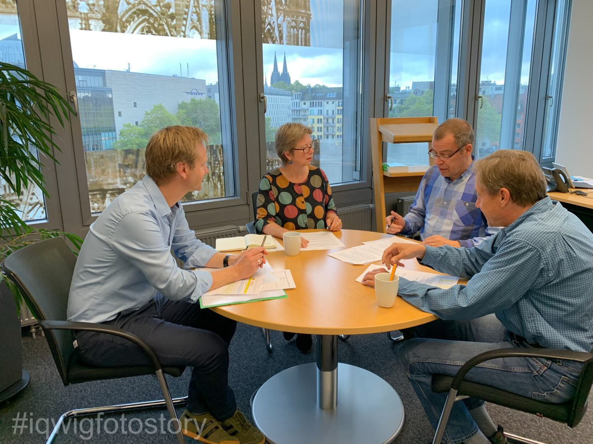 Three men in blue shirts and a woman in a top with large coloured dots are sitting around a round light-brown table and are discussing a text that is printed out in front of them. Through the window, you can see Cologne Cathedral in the distance.