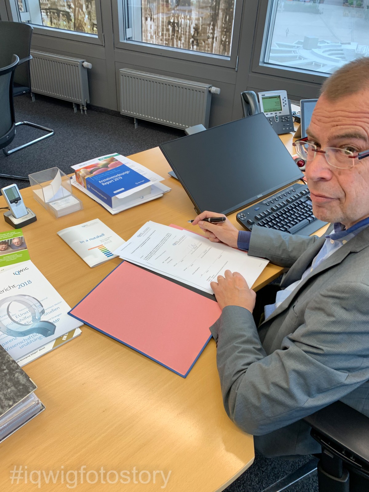 A man is sitting at a desk, looking at the camera. He has short hair, and is wearing glasses and a grey jacket with a light blue shirt underneath. There are documents in front of him. He is holding a pen in his right hand. On the table are books, pamphlets, a computer keyboard, a screen and a telephone. There are windows in the background to the right.