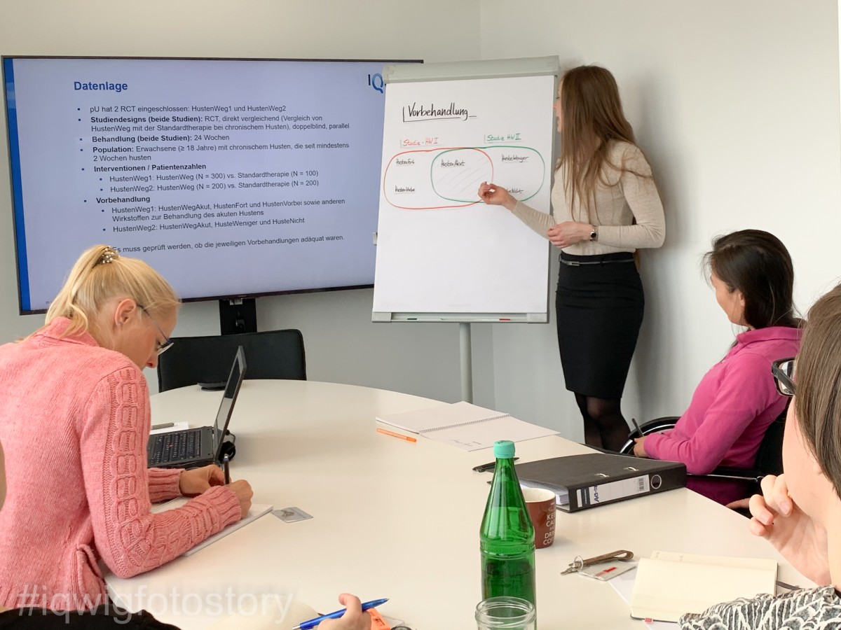 A woman is standing at a flipchart giving a presentation. People are seated around an oval table, intently following the presentation. To the left of the flipchart there is a large screen. On the table are a laptop, documents, notepads, a cup and a bottle of water.